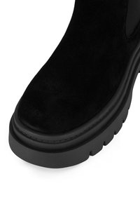 Czarny zamszowy botek za kostkę z zaokrąglonym noskiem, grubą czarną podeszwą i subtelnym przeszyciem wzdłuż krawędzi. Gładka faktura.