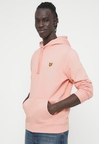 Ροζ φούτερ με κουκούλα από απαλό ύφασμα, με τσέπη καγκουρό και μικρό κίτρινο λογότυπο στο στήθος. Μακριά μανίκια με ριμπ μανσέτες.