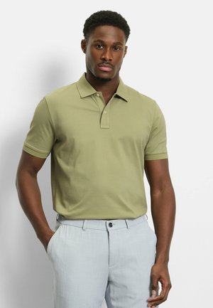 Mann trägt ein hellgrünes Polo-Shirt mit kurzen Ärmeln und eine hellgraue Hose, steht mit einer Hand in der Tasche vor einem einfarbigen Hintergrund.