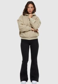 Giacca a cappuccio beige in pile con zip frontale, abbinata a jeans neri a zampa d'elefante e sneakers bianche; modella in piedi con le braccia incrociate.