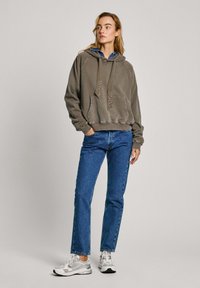 Sudadera con capucha color caqui y bolsillo frontal, decorada con tachuelas, combinada con jeans azules de pierna recta y zapatillas deportivas blancas.