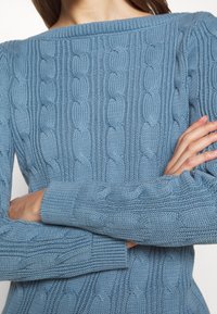 Personne portant un pull en maille torsadée bleu clair, les bras croisés, montrant la texture et les poignets côtelés sur un fond neutre.