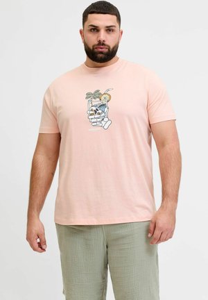 Szakállas férfi barackszínű pólóban, melyen egy csontváz kéz tart egy italt, valamint világoszöld texturált rövidnadrágot visel, fehér háttér előtt áll.