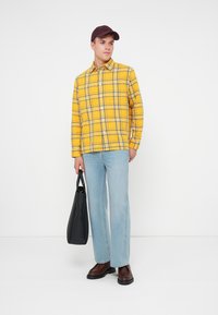 Gelb kariertes Langarmhemd, hellblaue gerade Jeans, braune Schuhe und dunkelblappe Mütze. Hält eine schwarze strukturierte Tragetasche.