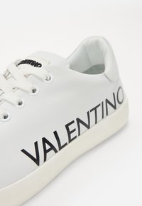 Bijela kožna tenisica s crnim natpisom "VALENTINO", okrugli vrh, dizajn s vezicama i gumeni potplat. Ima glatku teksturu i minimalne akcentne detalje.