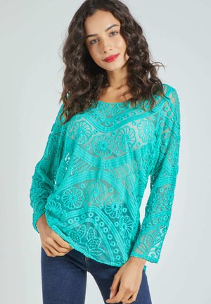 Una mujer con cabello rizado y oscuro lleva una blusa de encaje color turquesa brillante con patrones florales y jeans oscuros, posando frente a un fondo claro y liso.