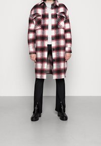 Chemise à carreaux en tissu rouge, noir et blanc ; conception à boutons avec de grandes poches poitrine. Portée par-dessus un pull gris clair et un pantalon noir avec des zips.