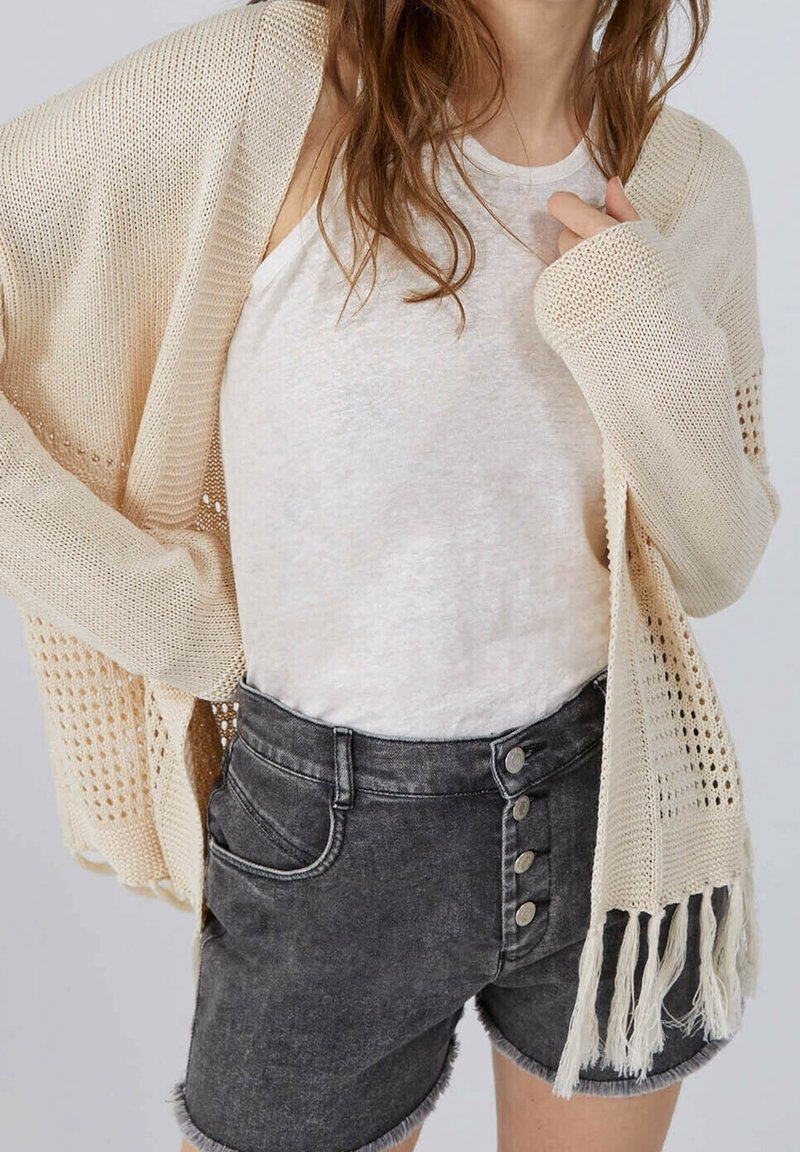 Béžový kardigan s otevřeným pletením a dlouhými rukávy, doplněný světle šedým topem a černými džínovými šortkami s vysokým pasem a knoflíkovými detaily.