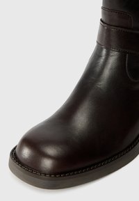 Dunkelbraune Lederstiefeletten mit runder Zehenform, glatter Oberfläche und zwei Riemen am Knöchel. Gestickte Details entlang der Sohle.