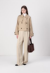 Cappotto trench cropped beige con ampi rever e bottoni marroni, abbinato a pantaloni larghi color crema e una borsa tote marrone scuro.