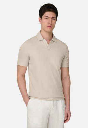 Jeune homme aux cheveux courts et foncés, portant un polo beige à manches courtes et un pantalon blanc, debout avec une main dans la poche.