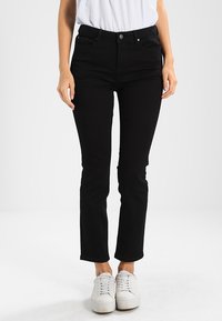 Vaqueros de denim negro con un corte slim, que cuentan con cinco bolsillos y cierre de botón. Llevados con zapatillas blancas, mostrando un diseño limpio.