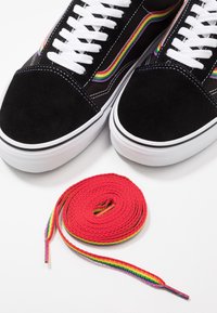 Černé tenisky s bílými podrážkami a šňůrkami, obohacené duhovým pruhem, vedle svinuté červené šňůrky s duhovými okraji na bílém povrchu.