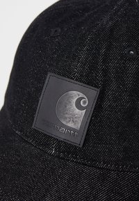 Juodas džinsinis kepurė su tekstūruota medžiaga, lenktu kraštu ir stačiakampiu guminiu logotipo ženkleliu, kuriame pavaizduota Carhartt emblema.
