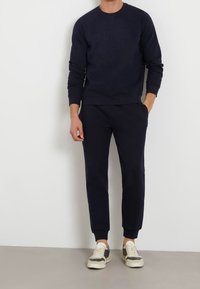 Man som bär en marinblå sweatshirt och matchande joggingsbyxor med vita och grå sneakers, står mot en enkel vit vägg.