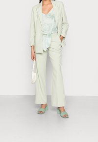 Miętowozielony garnitur damski z bluzką z wiązaniem z przodu, dopasowaną marynarką i miękkim materiałem. Do zestawu dobrano zielone sandały na masywnym obcasie oraz białą torebkę.
