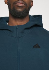 Bearded person wearing a zipped-up dark teal Adidas hoodie with a hood and black logo on the chest against a plain background.