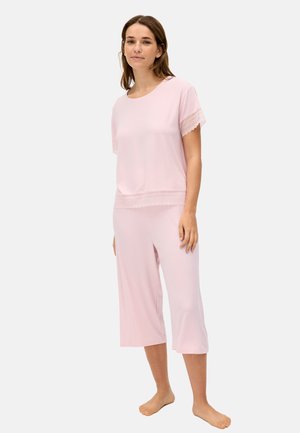 Mujer con camiseta rosa claro de manga corta y pantalones recortados con encaje, de pie descalza contra un fondo liso.