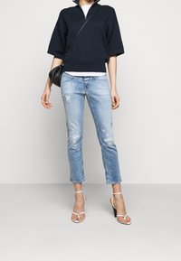 Persona que lleva unos vaqueros cortos de mezclilla azul claro desgastados, una camiseta navy de manga corta, unos zapatos de tacón blancos con tiras y un pequeño bolso negro cruzado.