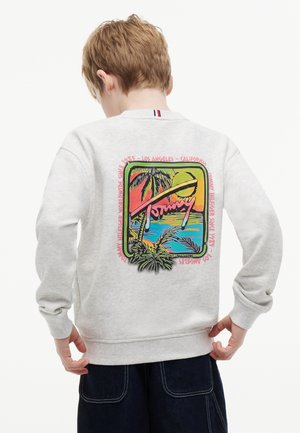 Dziecko z krótkimi jasnobrązowymi włosami, ubrane w jasnoszarą bluzę z kolorową grafiką tropikalnego zachodu słońca i palmy na plecach.