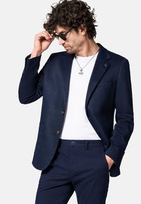 Granatowa marynarka na dwa guziki, z klapami w kształcie notched i kieszeniami z przodu, zestawiona z białą koszulą i granatowymi spodniami. Strukturalny materiał i metaliczne akcenty.