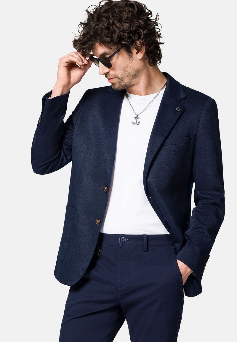 Granatowa marynarka na dwa guziki, z klapami w kształcie notched i kieszeniami z przodu, zestawiona z białą koszulą i granatowymi spodniami. Strukturalny materiał i metaliczne akcenty.