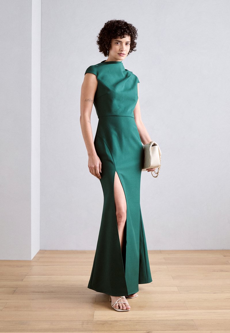 Zielona, dopasowana wieczorowa suknia z rękawami typu cap oraz rozcięciem na udzie; wykonana z gładkiego materiału; zestawiona z torebką w kolorze kremowym i sandałami.