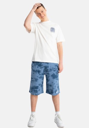 Młody mężczyzna w białym T-shircie z grafiką i niebieskich szortach tie-dye, stoi z jedną ręką na głowie i białych tenisówkach na białym tle.