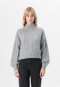 Blonde Frau in einem grauen, kabelstrickmuster Pullover mit hohem Kragen und langen Puffärmeln sowie schwarzen, hoch taillierten Hosen vor weißem Hintergrund.