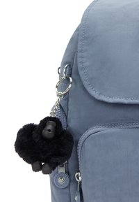 Grå tygryggsäck med klaffstängning, med en svart fluffig gorillanyckelring. Dragkedjor och hållbara sömmar synliga.