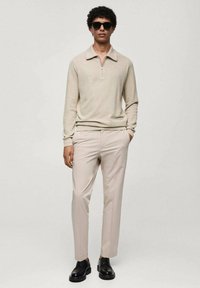 Beige zip-hals trøje, lysebeige skræddersyede bukser og sorte snørebåndssko. Model iført sorte solbriller, står med hånden i lommen.