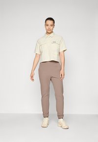 Lichtbeige cropped shirt met twee voorzakken, gecombineerd met strakke bruine broeken. Lichtbeige sneakers maken de look compleet.