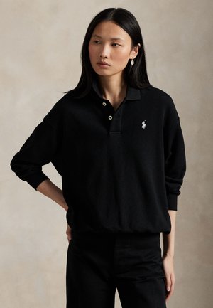 Femme aux longs cheveux foncés portant un polo noir à manches longues avec un petit logo blanc et un pantalon noir, debout sur un fond uni.