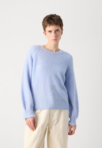 Lys blå ullgenser med rund hals, ribbestrikkede mansjetter og en litt oversized passform. Har en liten sølvdetalj på siden.