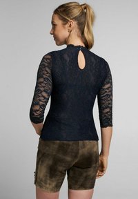 Marineblaues Spitzenoberteil mit Dreiviertelärmeln, Rückendetails mit Schlüsselloch-Ausschnitt, gewellten Kanten, kombiniert mit braunen Wildleder-Shorts aus strukturiertem Stoff.