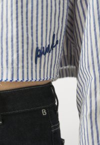 Camicia a righe blu e bianche con logo ricamato "pink" sull'orlo. Presenta un design cropped con bordi sfrangiati. Abbinata a jeans scuri.