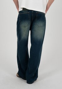 Persoană care poartă blugi largi din denim albastru închis, cu zone decolorate pe buzunarele din spate și pe coapse, stând în fața unui fundal simplu, deschis la culoare.