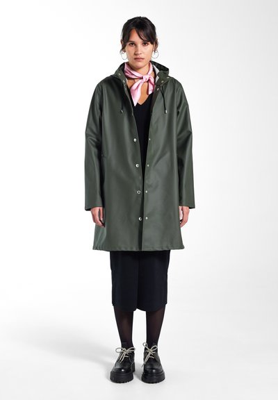 Imperméable vert, résistant à l'eau, avec capuche, boutons pression et poches latérales ; associé à une robe noire et des chaussures noires imposantes. Écharpe rose en accent.