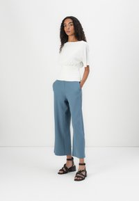 Witte top met korte mouwen en een gerimpelde taille, gecombineerd met lichtblauwe wijde broek en zwarte haksandalen. Minimalistisch ontwerp en gladde textuur.