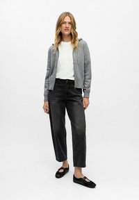 Kobieta z długimi blond włosami, ubrana w szary rozpinany hoodie, białą koszulę, czarne dżinsy 3/4 i czarne buty wsuwane, stojąca na jednolitym tle.