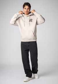 Lichtgrijs hooded sweatshirt met een voorzak en een grafisch ontwerp op de borst. Gecombineerd met zwarte jeans en witte sneakers.