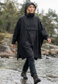 Femme portant un imperméable noir à capuche marchant sur un terrain rocheux humide près de l'eau et des arbres sous une pluie légère.