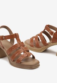 Sandales à talons en cuir marron avec plusieurs lanières et un design tressé. Comprend une fermeture à boucle et une semelle plateforme courbée de couleur beige.