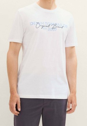 Mand iført en hvid T-shirt med korte ærmer med blå og sort tekst og mørke bukser, står mod en ensfarvet lys baggrund.