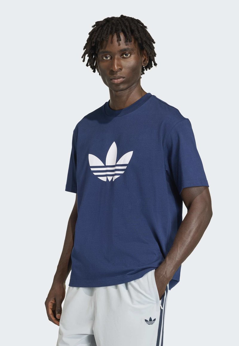Uomo con dreadlocks che indossa una maglietta Adidas blu navy e pantaloni della tuta Adidas bianchi, in piedi con una mano in tasca.