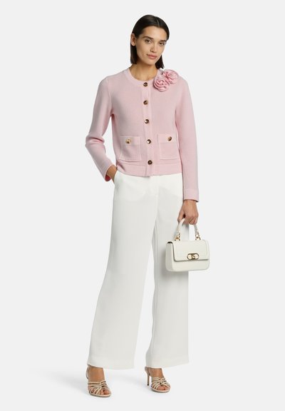 Svetlo roza pletena kardigan s prednjo detajlno rožo, zlatimi gumbi in dvema žepoma, v kombinaciji z belimi širokimi hlačami in majhno belo torbico.