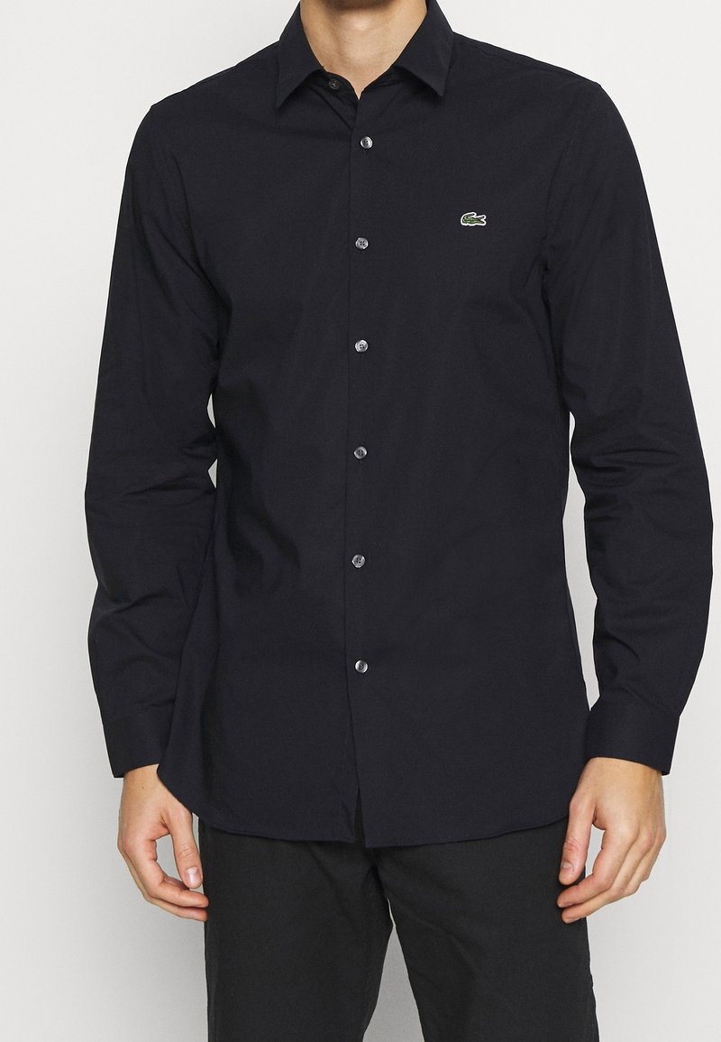 Black long-sleeve shirt with a pointed collar, button front, and a small green logo on the chest. Smooth texture, tailored fit, and solid color.