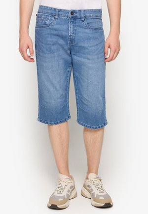 Mand iført midt-lange blå denimshorts med sømdetaljer og hvide beige sneakers, der står med fronten mod kameraet.