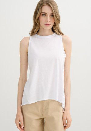 Joven mujer con cabello claro que lleva una camiseta blanca sin mangas y pantalones beige, de pie frente a un fondo claro y liso.