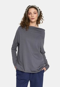 Kobieta z kręconymi włosami, ubrana w szary, długi rękaw, niebieskie spodnie oraz szary opaska na głowie, stojąca z rękami w kieszeniach na białym tle.